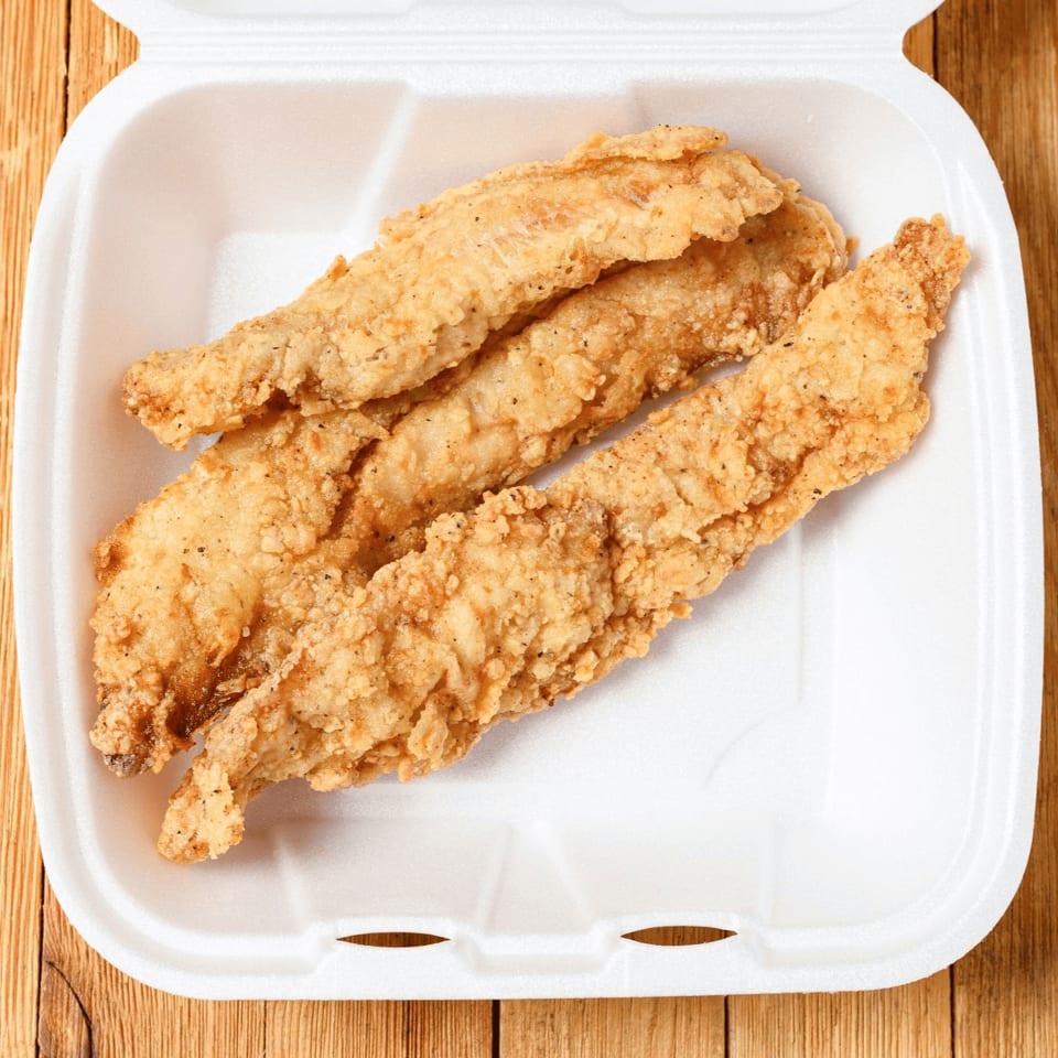 Three golden-brown fried chicken tenders in a white styrofoam takeout container on a wooden surface.