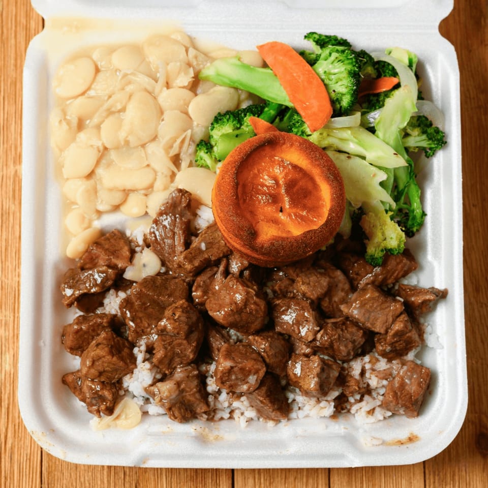 A takeout container filled with beef tips over rice, lima beans, mixed vegetables, and a cornbread muffin.