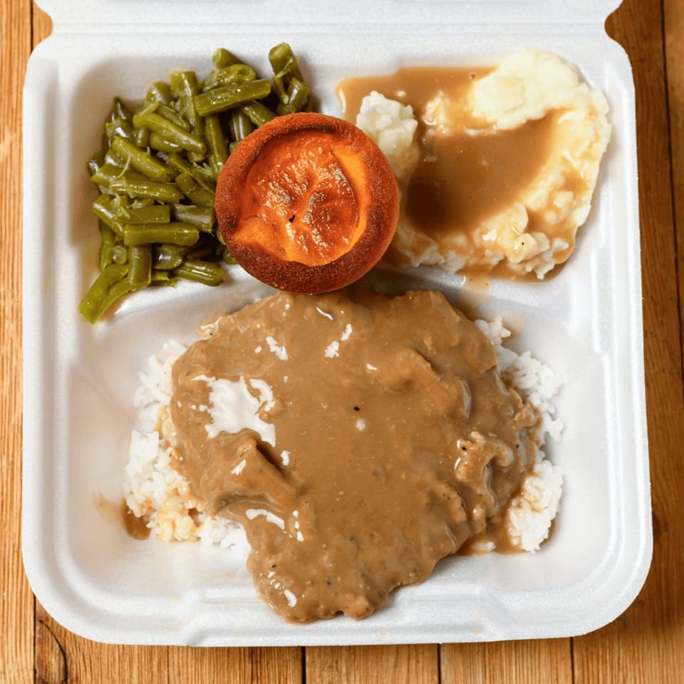 A takeout container with Salisbury steak, rice, mashed potatoes, gravy, green beans, and a roll.