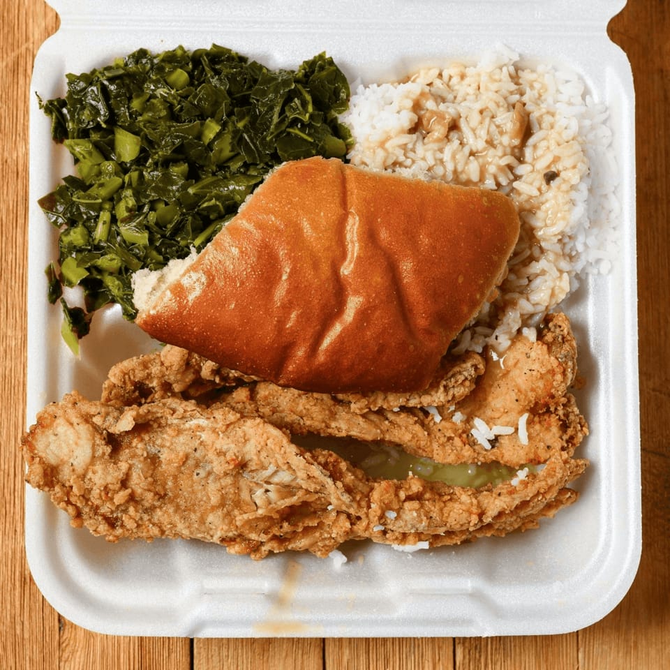 Fried chicken, collard greens, rice with gravy, and a roll in a white takeout container.
